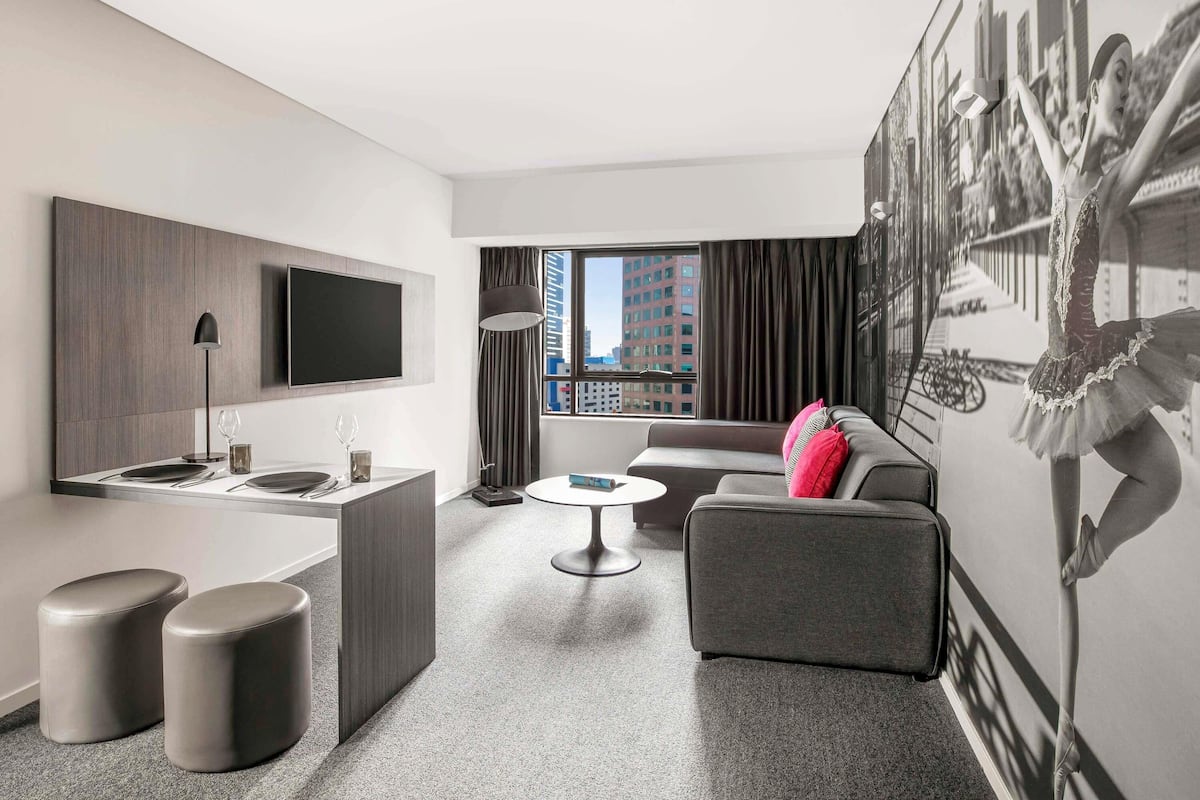 A modern hotel room with a flat-screen TV, a dining area, a sofa, and a large window with a city view.