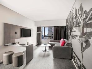 Ein modernes Hotelzimmer mit einem Flachbildfernseher, einem Essbereich, einer Couch und einem großen Fenster mit Blick auf die Stadt.