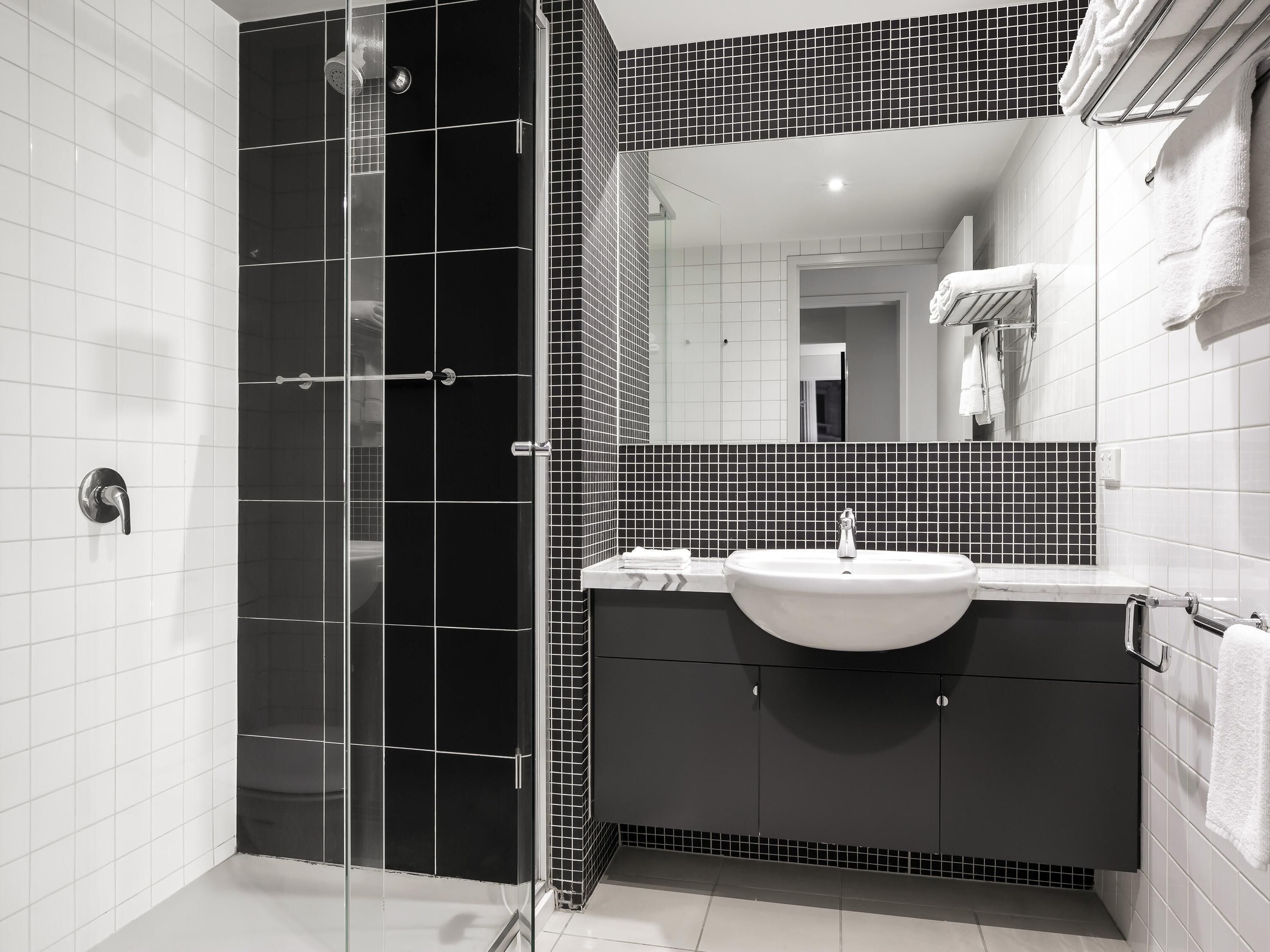 Une salle de bain moderne avec une douche à l’italienne en verre, un lavabo blanc, des meubles aux tons foncés et un mur recouvert de carreaux.