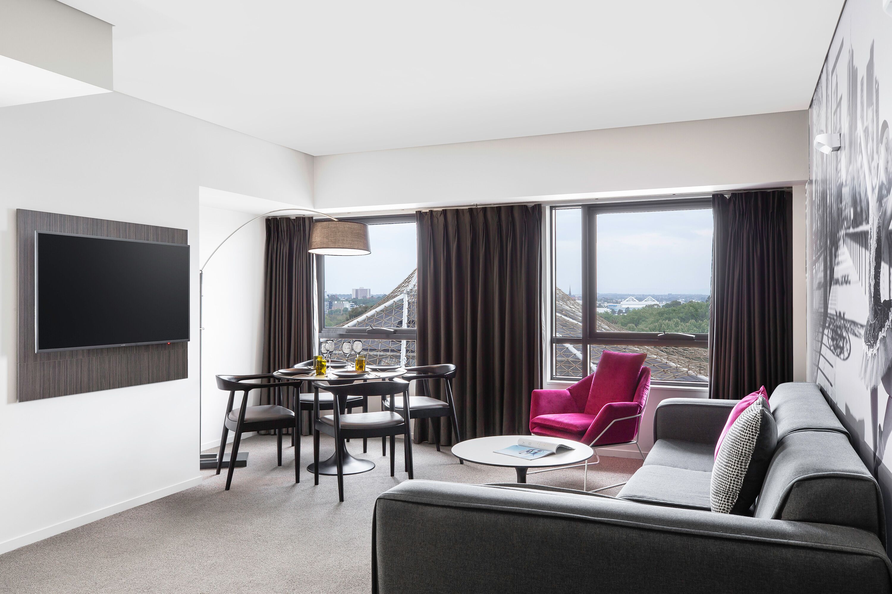 Een moderne hotelkamer met een flatscreen televisie aan de muur, een zithoek met een tafel en stoelen, een grijze bank, een rode fauteuil en grote ramen met uitzicht op de stad.
