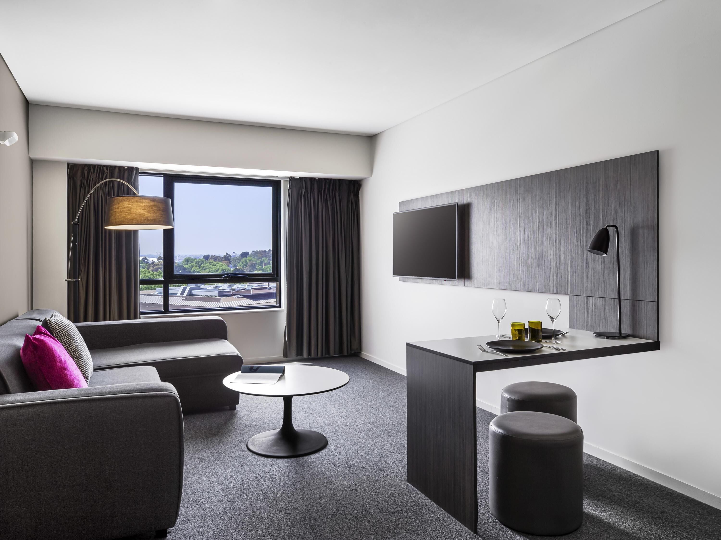 Una habitación de hotel moderna con un sofá, una mesita redonda, un televisor montado en la pared y una ventana con cortinas.