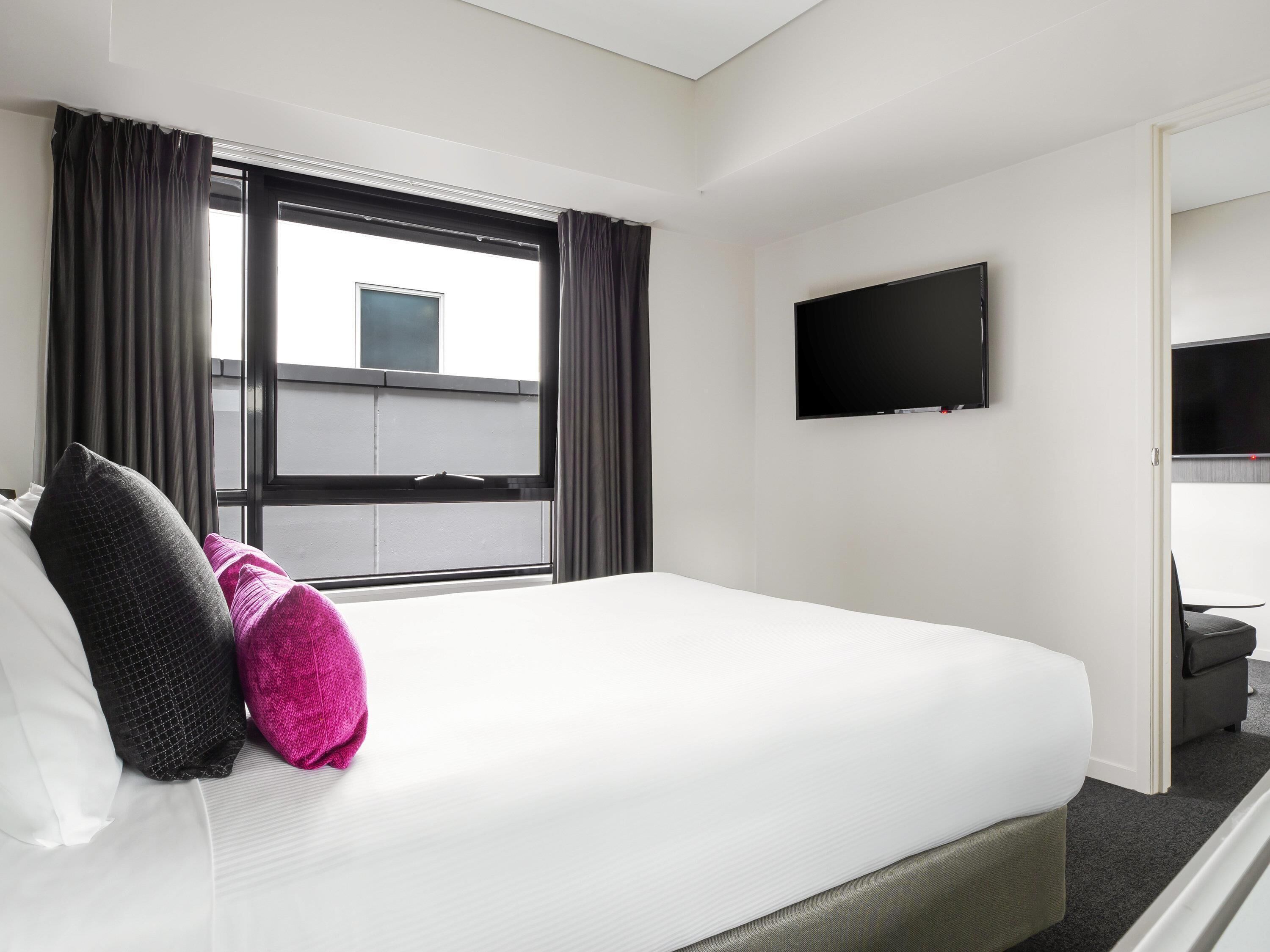 A hotel room with a large bed, a flat-screen TV mounted on the wall, and a window with curtains.