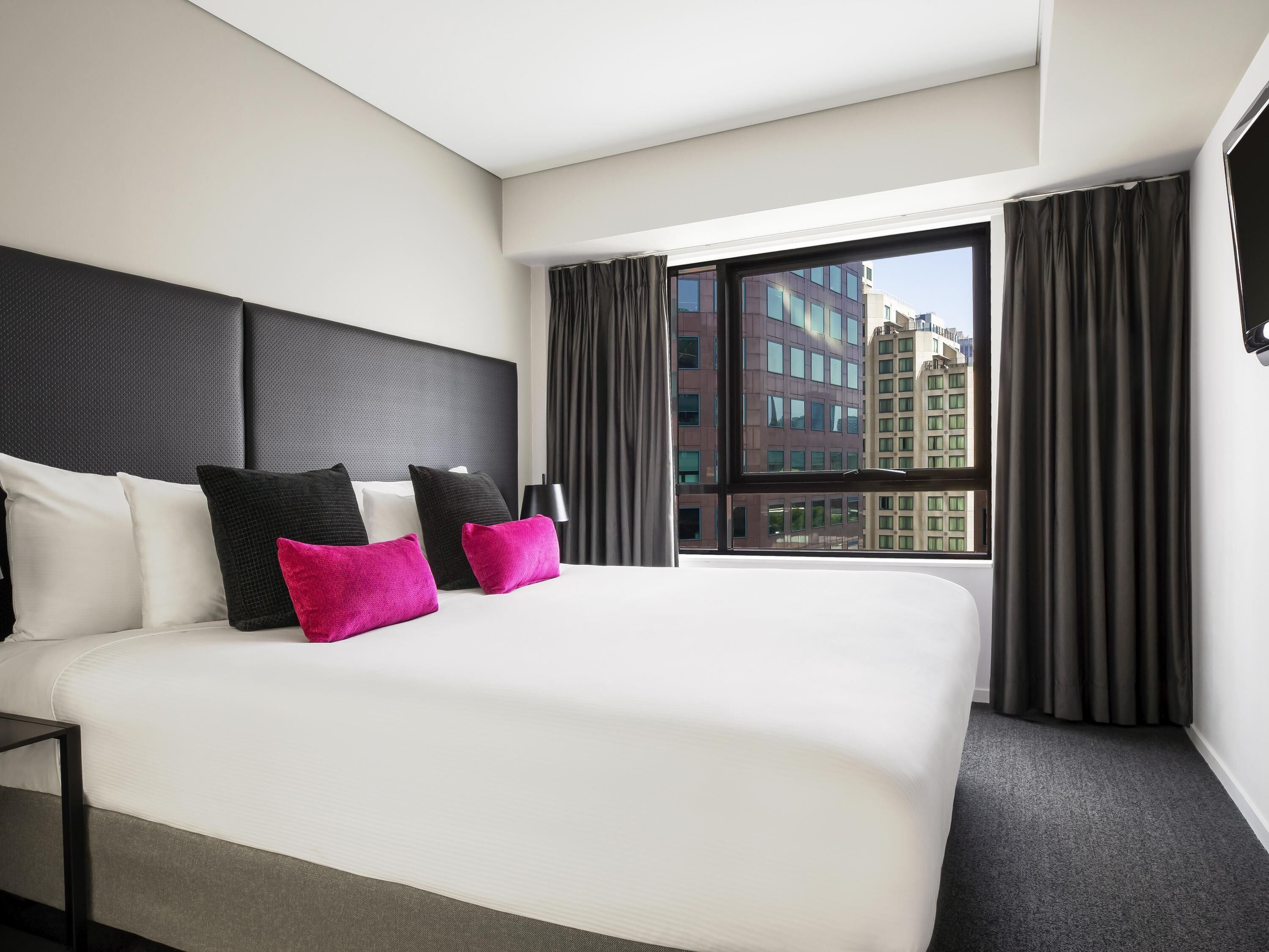 A hotel room with a large bed, a headboard, a window with curtains, and a view of buildings.