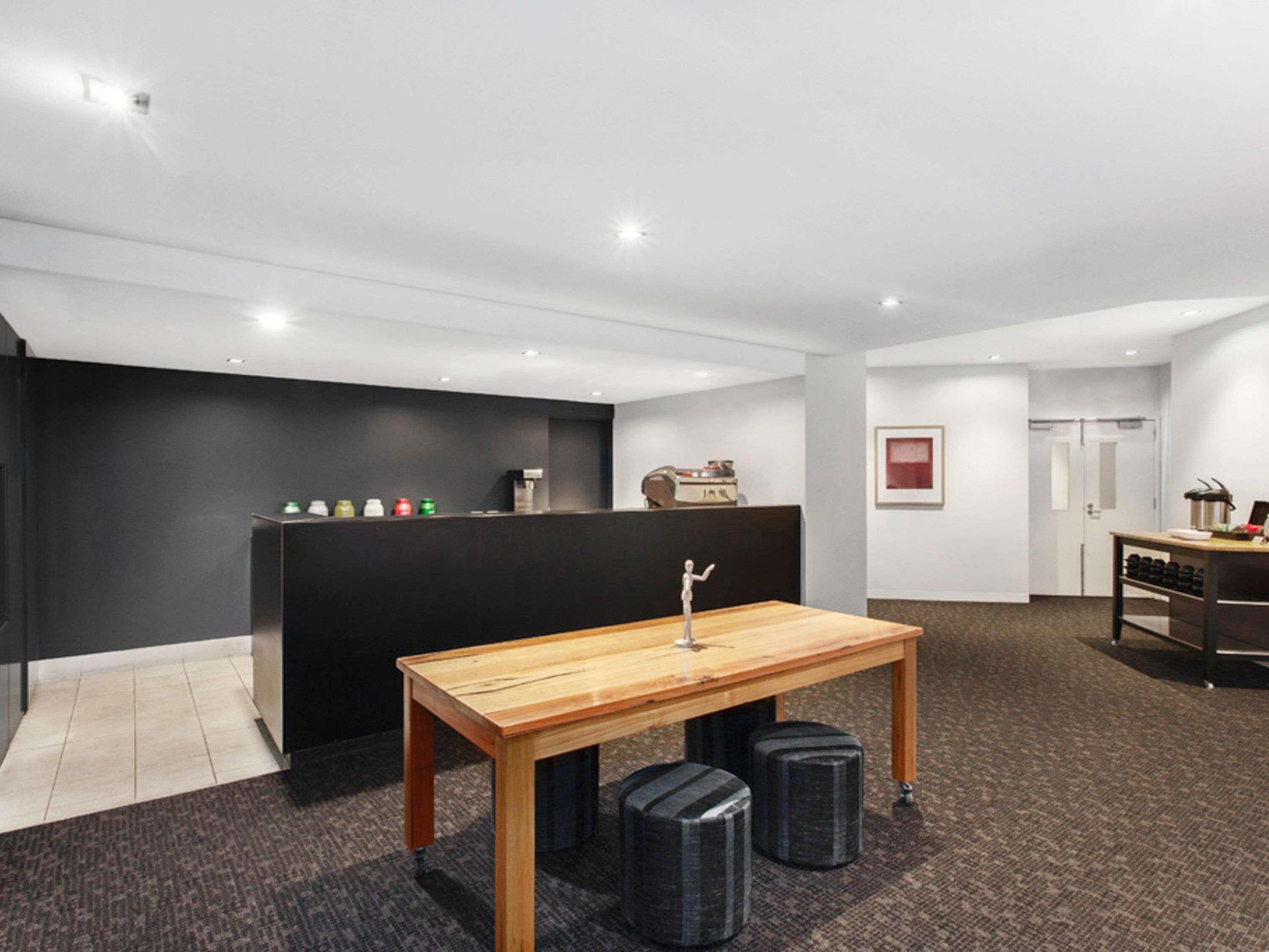 A modern hotel room with a wooden table, black ottomans, a reception desk, and a framed picture on the wall.
