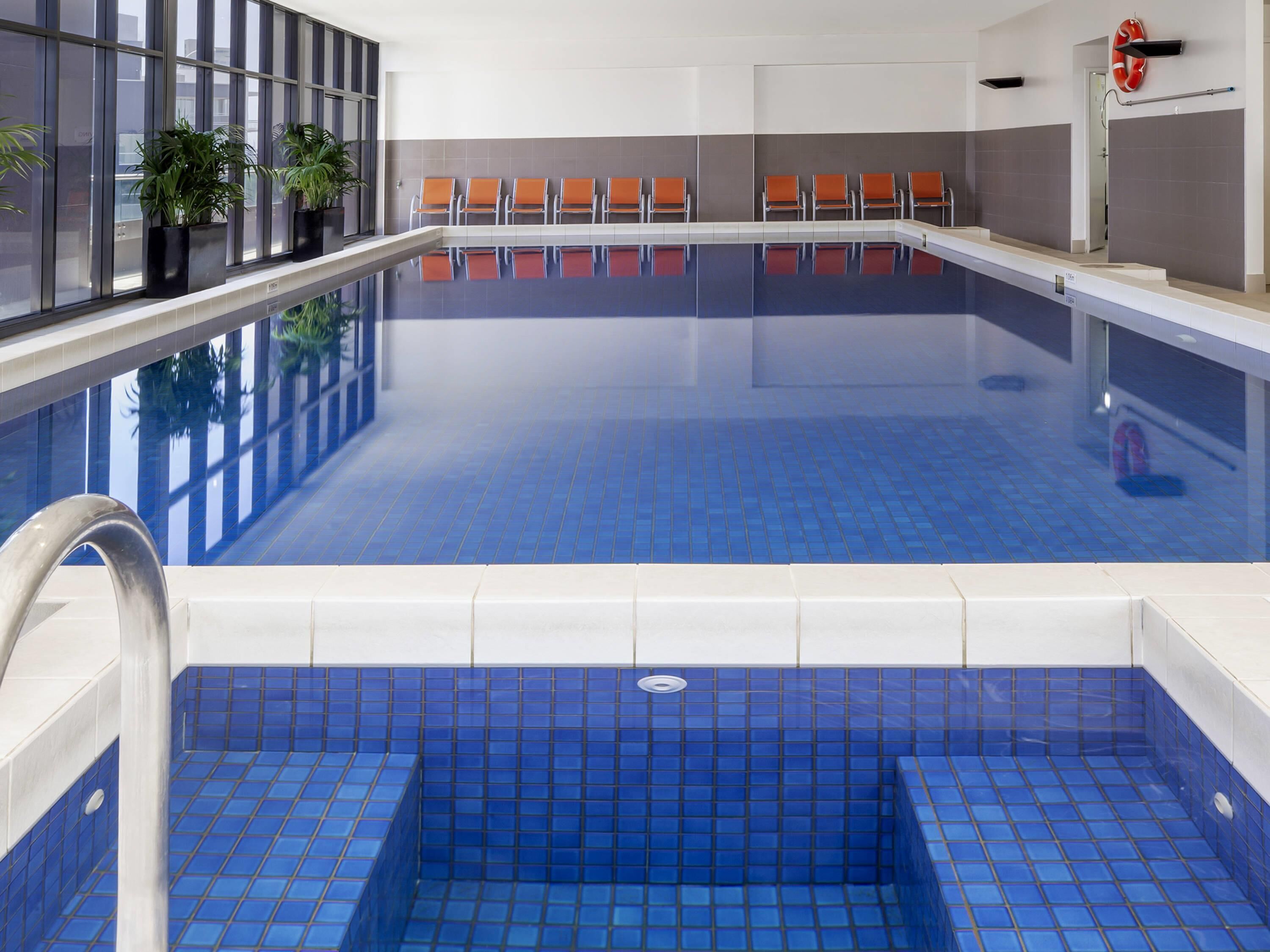 A swimming pool with blue tiles, a seating area with orange chairs, and potted plants along the window.
