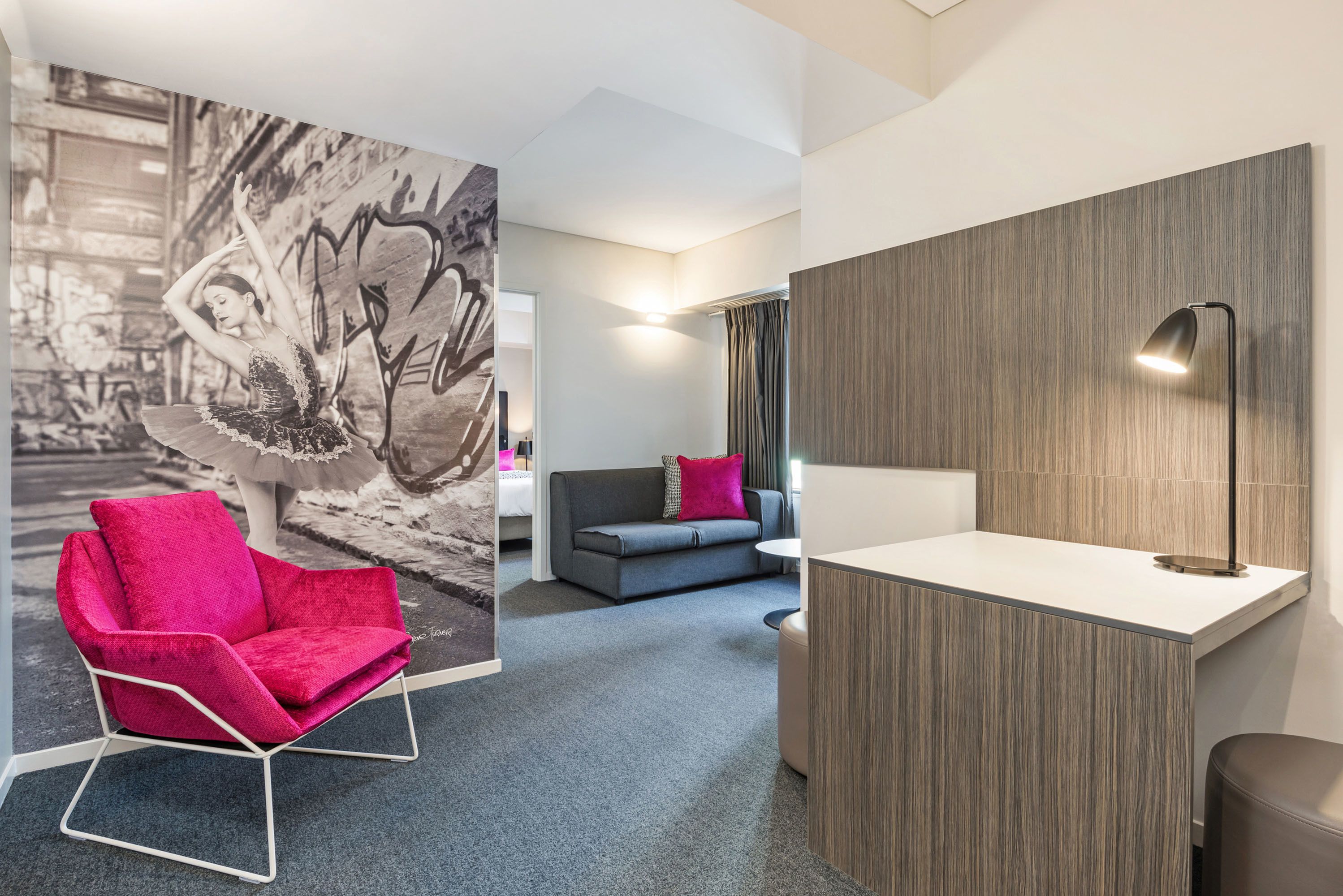 Una habitación de hotel moderna con un sofá gris, una silla roja, una pequeña mesa blanca y una lámpara fijada a la pared.