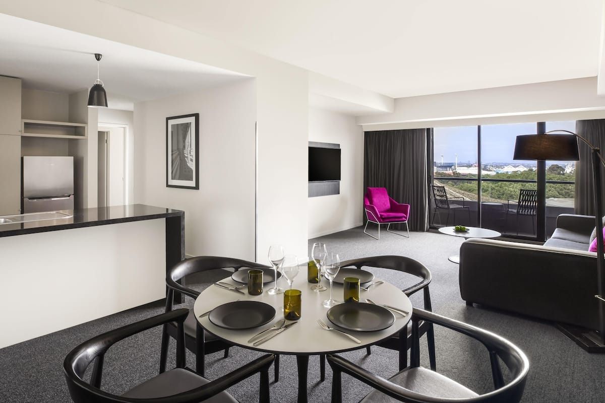 A modern hotel room with a dining area, a sofa, a balcony with a view, and a flat-screen TV.