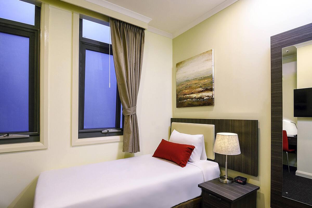 Ein Hotelzimmer mit Bett, Nachttisch, Lampe, einem Gemälde an der Wand und einem großen Spiegel.