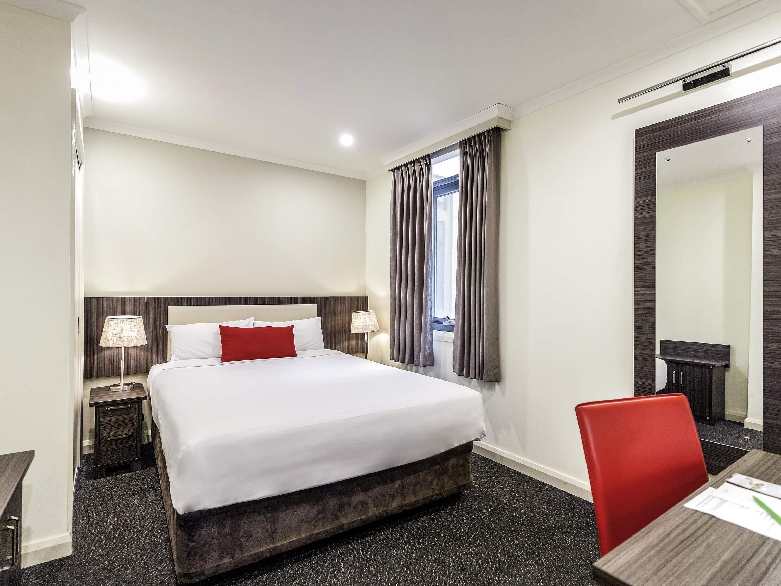 Habitación de hotel con cama, lámparas de noche, escritorio con silla, ventana con cortinas y un espejo.