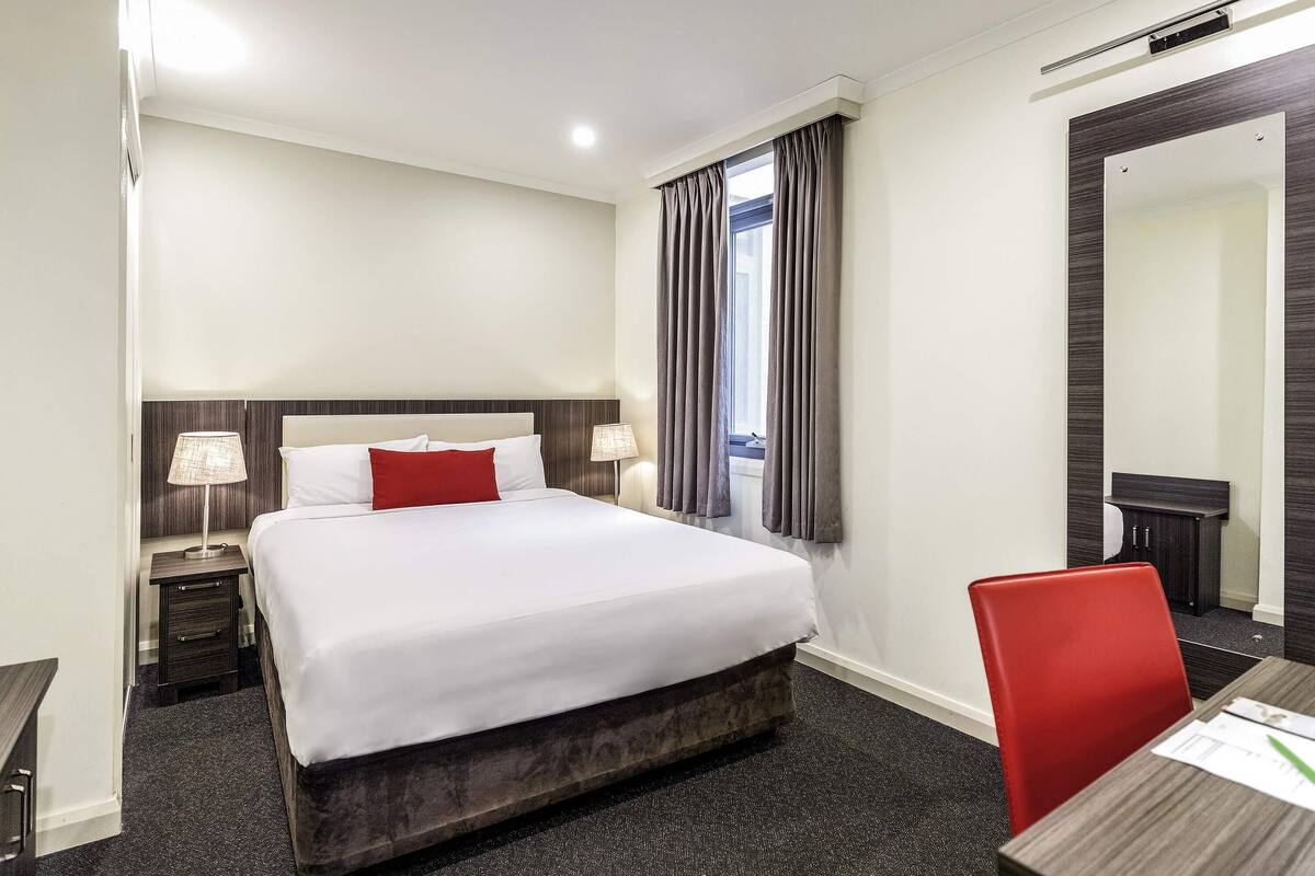 Habitación de hotel con cama, lámparas de noche, escritorio con silla, ventana con cortinas y un espejo.