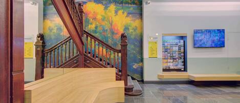 A staircase with wooden railings leading to a lower level, a mural with autumn foliage, a digital display, and a sign indicating amenities.
