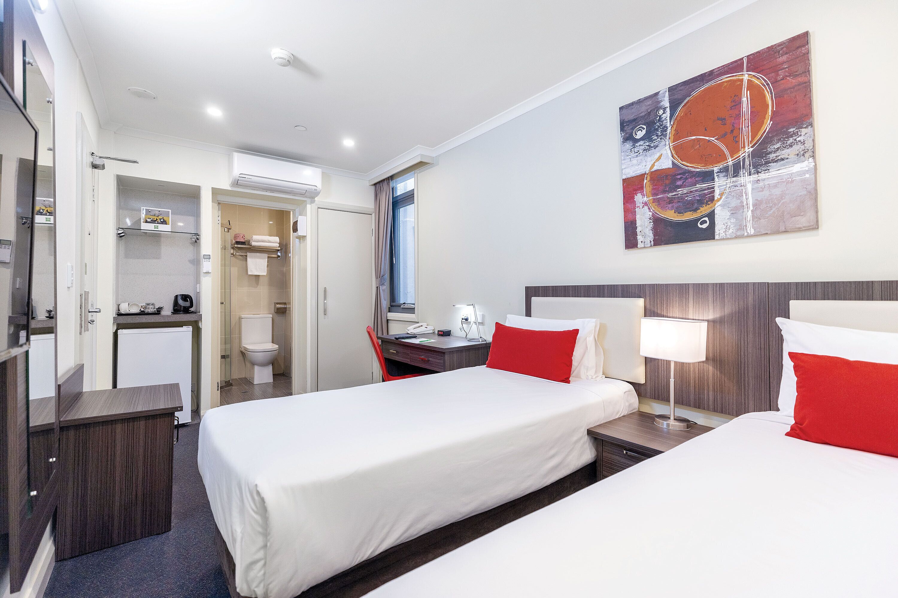 A hotel room with two beds, a desk, a chair, a small kitchenette, and a bathroom.
