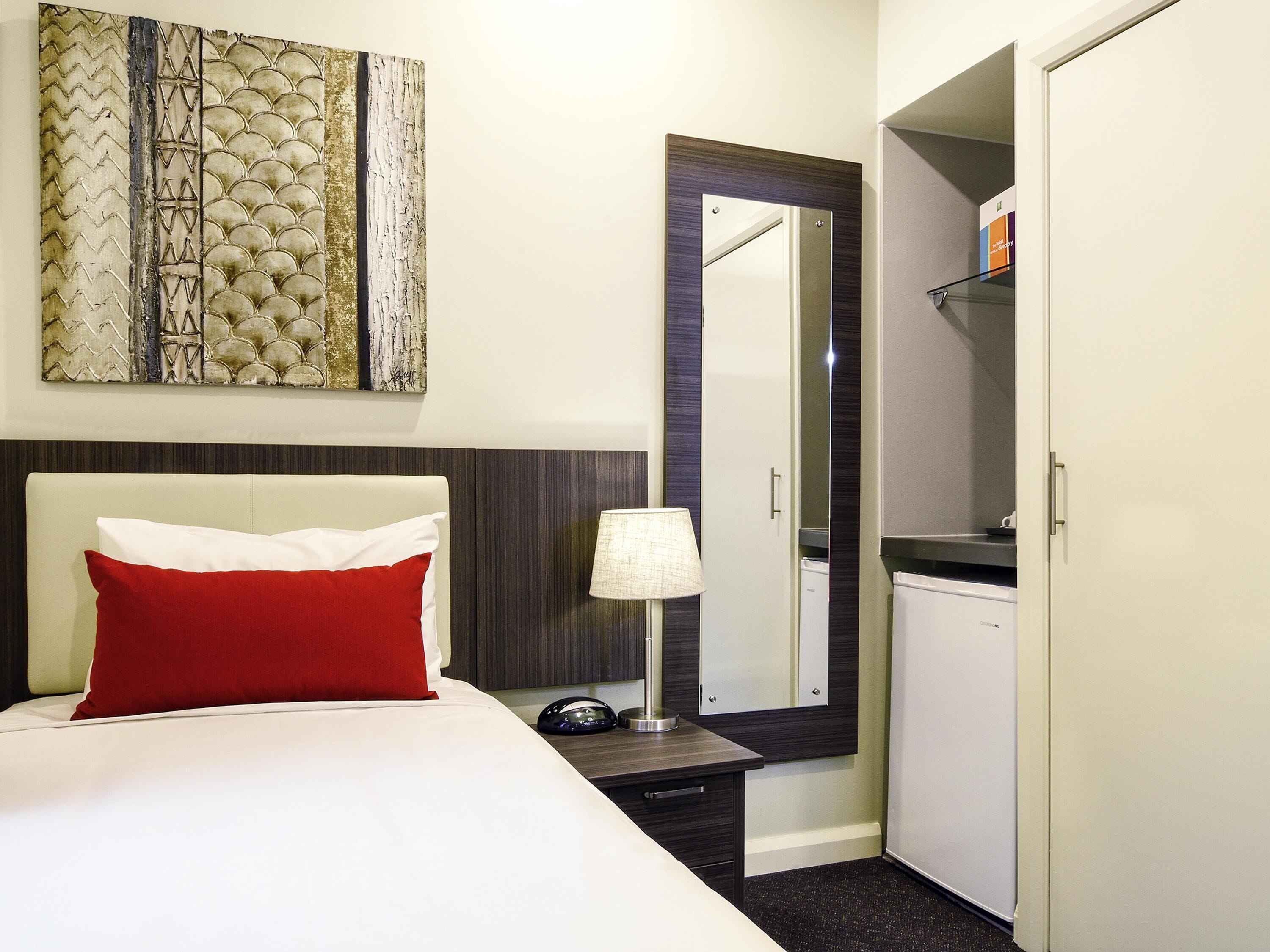 A hotel room with a bed, a nightstand, a lamp, a mirror, and a wardrobe.