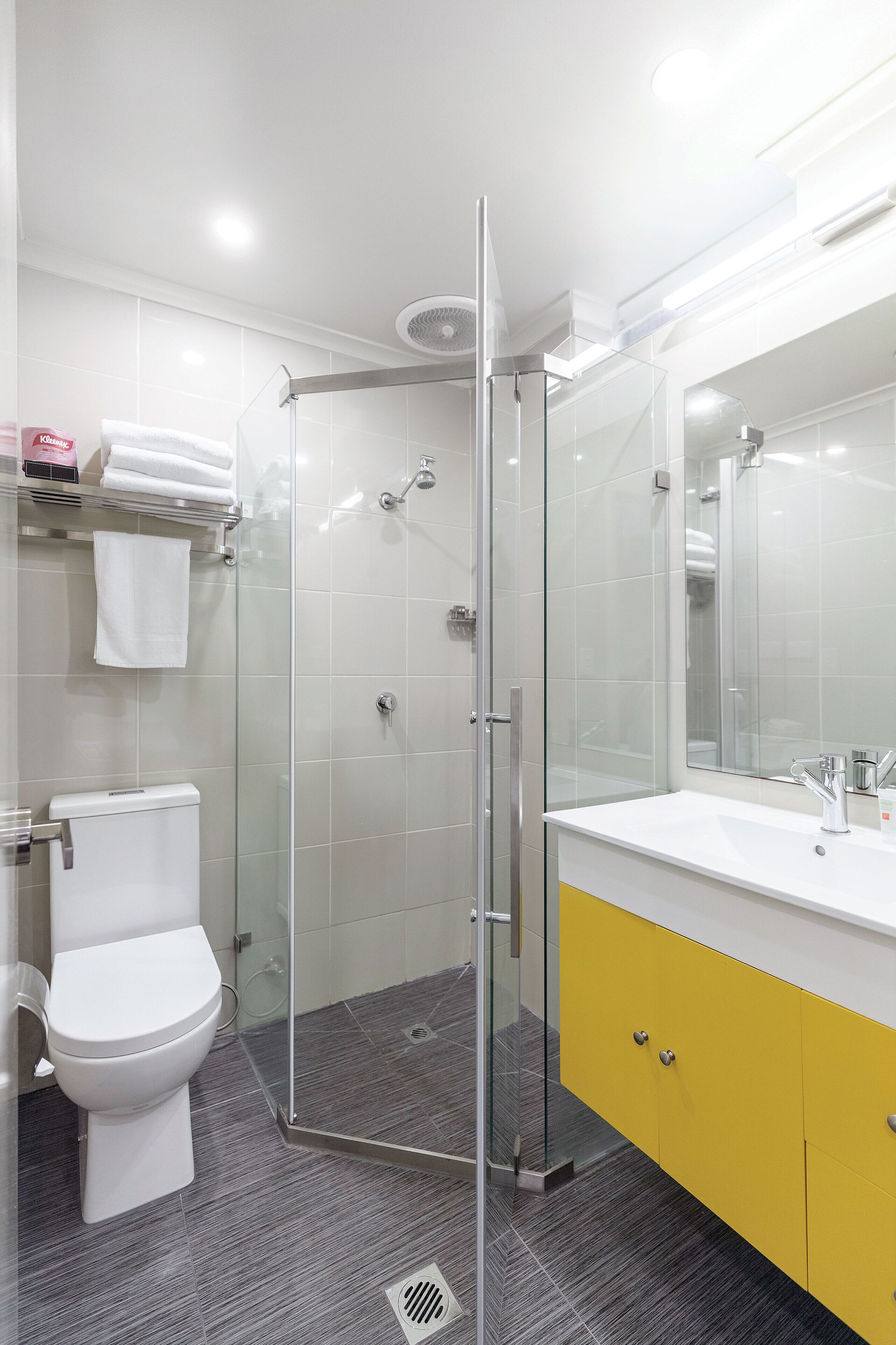 Une salle de bain moderne avec une douche à parois de verre, des toilettes blanches et un meuble-lavabo jaune agrémenté d’un grand miroir.