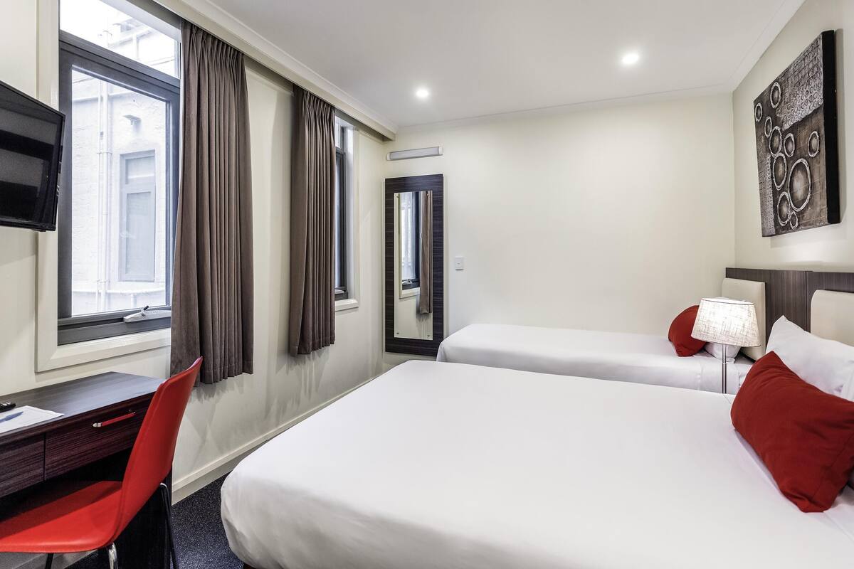 Ein Hotelzimmer mit zwei Betten, einem Schreibtisch, einem Stuhl, einem Fernseher und einem Fenster mit Vorhängen.