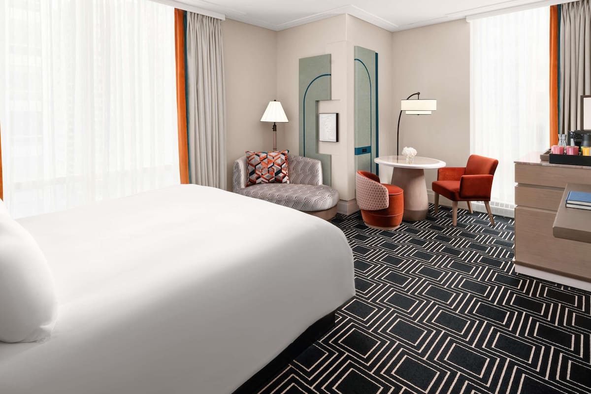 A hotel room with a large bed, a desk with a chair, a small table with a lamp, and a sofa with a cushion.