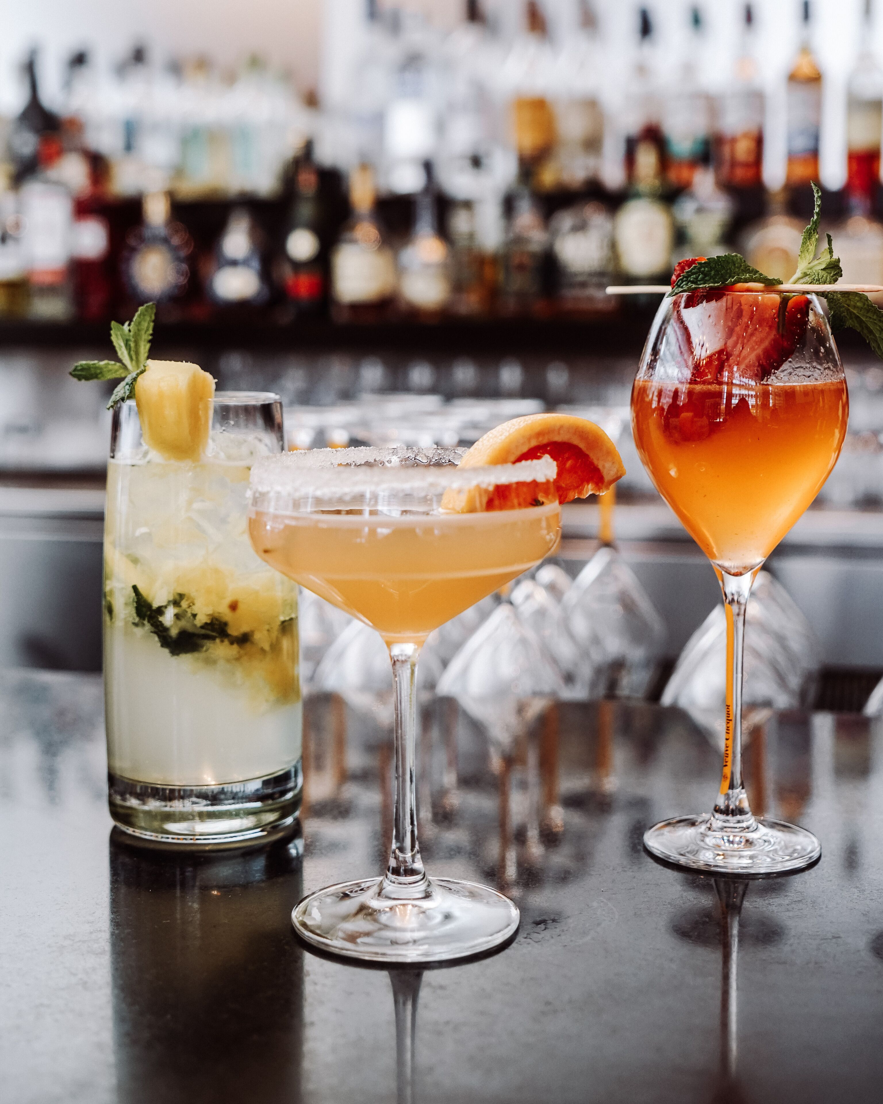 Three cocktails on a bar counter with a blurred background of bottles.