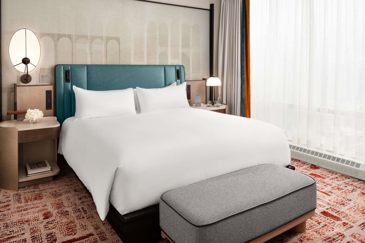A hotel room with a bed, bedside tables, a lamp, a small table with a vase, and a window with curtains.