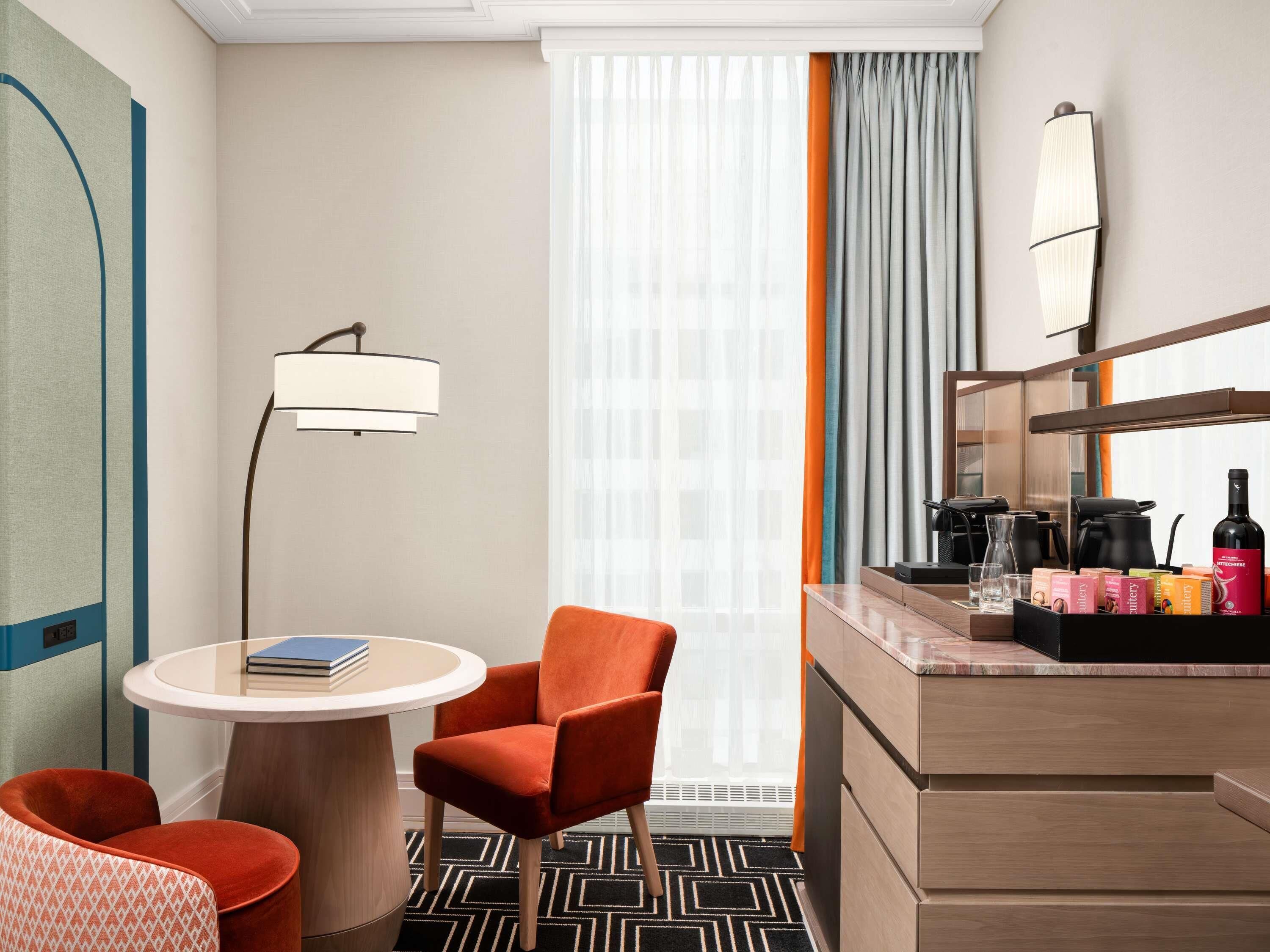 Habitación de hotel con una mesa redonda, dos sillas, un minibar, una cafetera y un ventanal grande con cortinas.