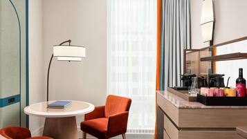 Habitación de hotel con una mesa redonda, dos sillas, un minibar, una cafetera y un ventanal grande con cortinas.