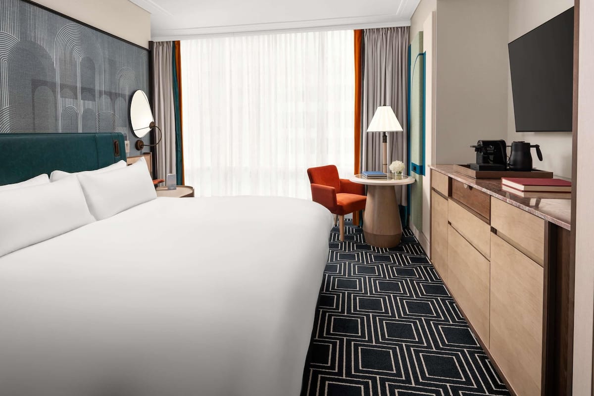 A hotel room with a large bed, a desk with a lamp, a chair, and a TV.
