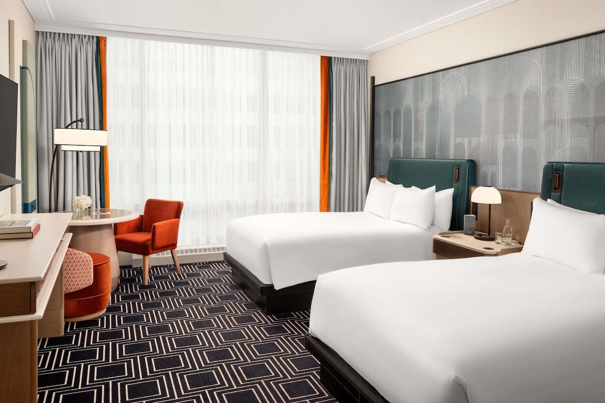 A hotel room with two beds, a desk, a chair, and a large window with curtains.