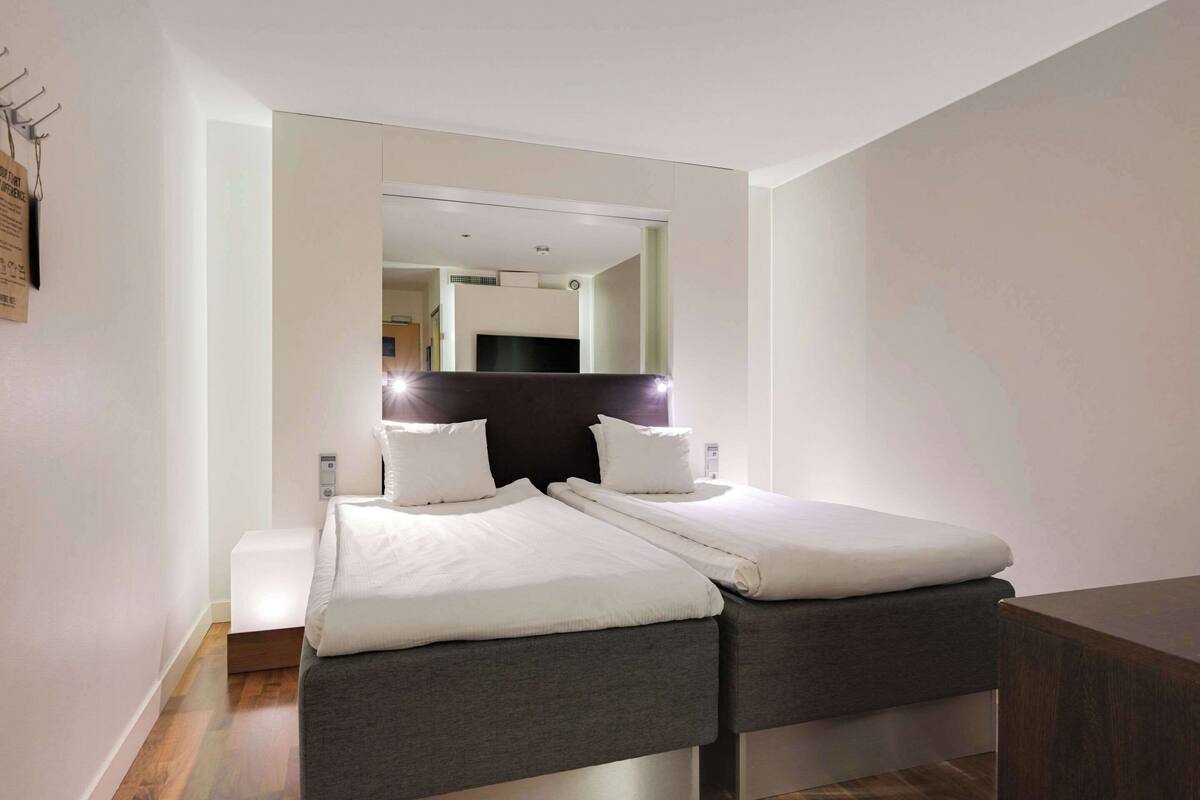 Una habitación de hotel moderna con una cama grande, una mesita de noche pequeña y un espejo sobre la cama.