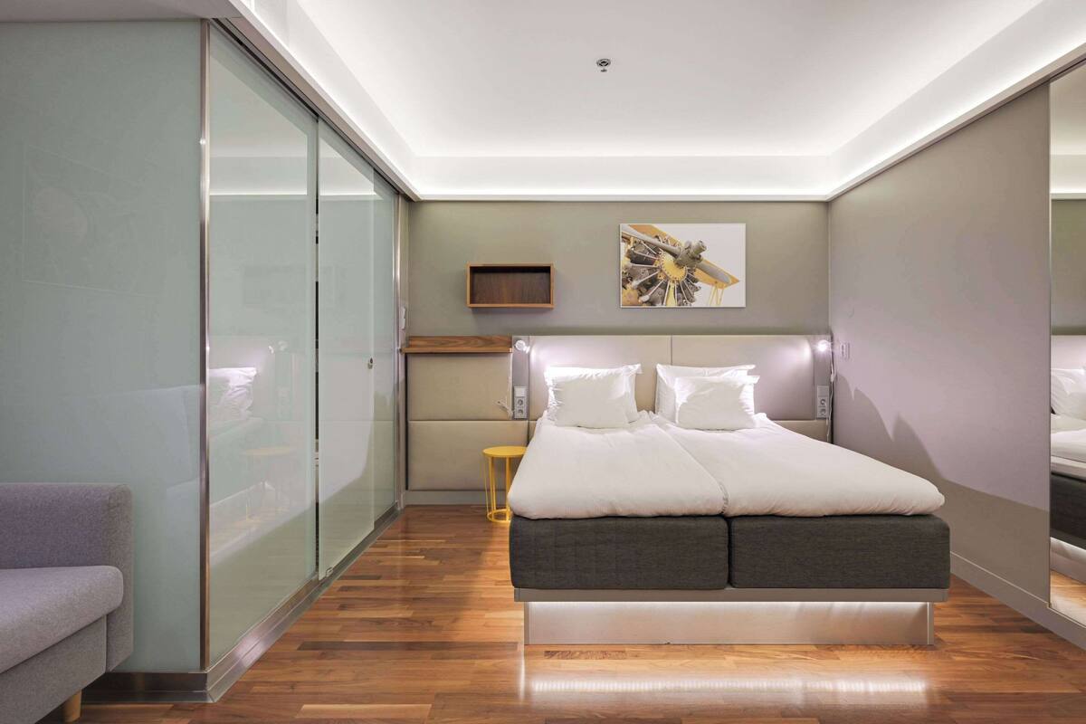 Un dormitorio moderno con una cama, un sofá, un separador de vidrio y un pequeño estante de madera.