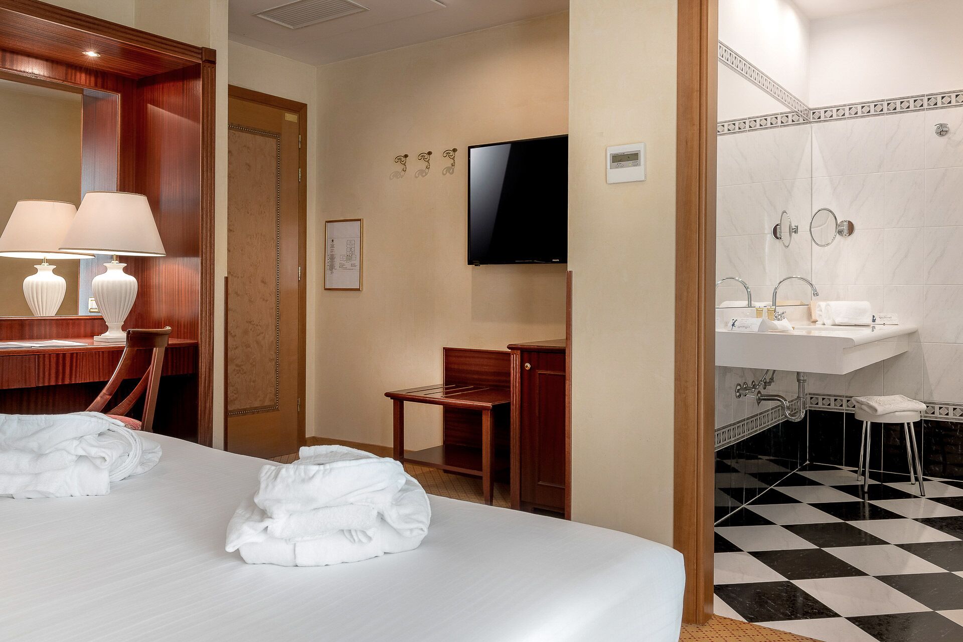 Habitación de hotel con cama, escritorio con lámpara, silla, televisor y baño con lavamanos y espejo.
