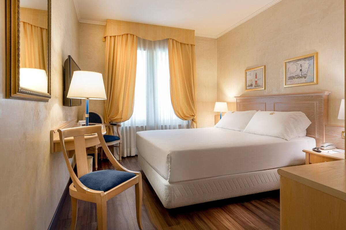 Ein Hotelzimmer mit Bett, zwei Nachttischen, einem Sessel, einem Schreibtisch und einem Spiegel.