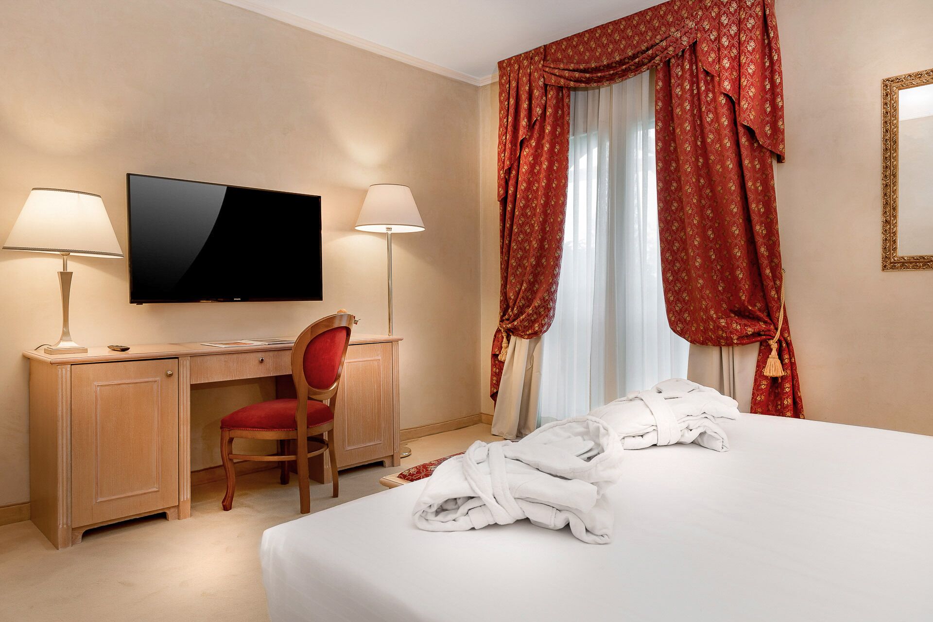 Quarto de hotel com cama, escrivaninha com cadeira, televisão e janela com cortinas.
