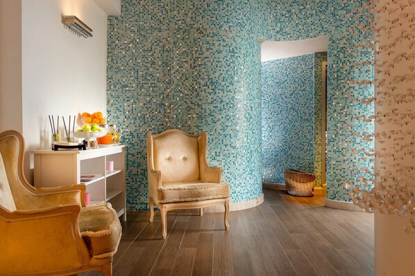 A room with a mosaic tiled wall, a wooden floor, a beige armchair, a small table with fruit and drinks, and a wicker basket. - Hotel De Londres (Rimini)