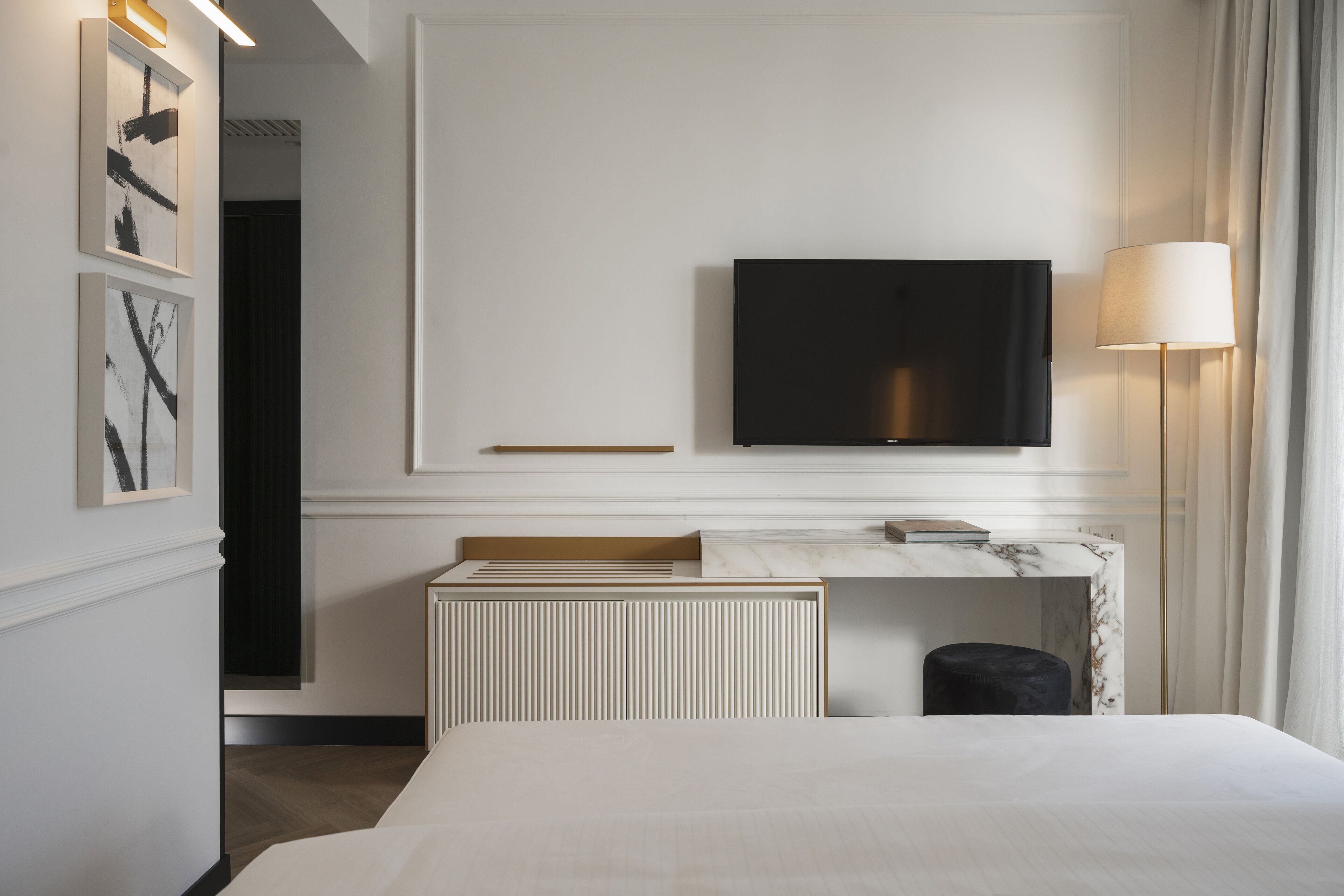 A hotel room with a large bed, a flat-screen TV mounted on the wall, a marble-topped desk, and a standing lamp.