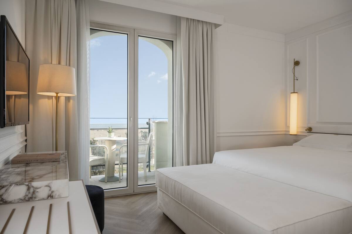 Ein Hotelzimmer mit einem großen Bett, einem Schreibtisch mit Lampe, einem Fernseher und Blick ins Freie durch eine Schiebetür.