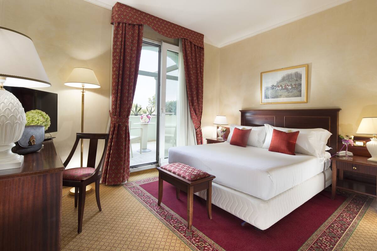 Ein Hotelzimmer mit einem großen Bett, einem Schreibtisch, einem Sessel, einem kleinen Tisch und einem Balkon mit Vorhängen.