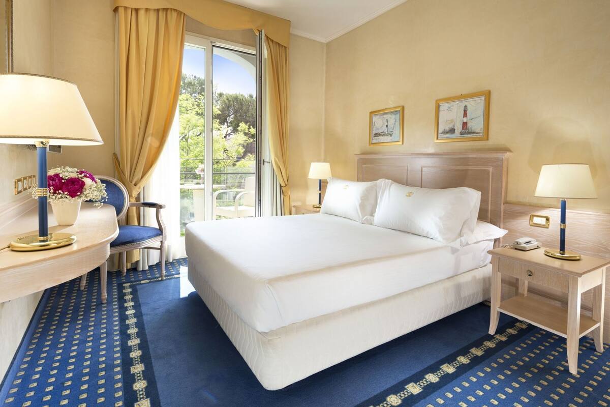 Ein Hotelzimmer mit einem großen Bett, zwei Nachttischen mit Lampen, einem Sessel, einem Schreibtisch mit Lampe und Blumen sowie Blick durch eine Glastür.