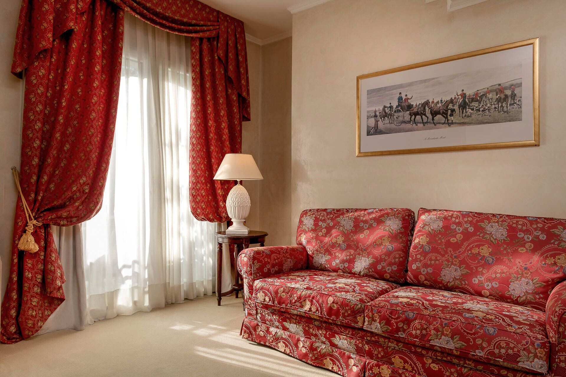 Una habitación con un sofá rojo estampado, un cuadro histórico enmarco y cortinas rojas con flecos.