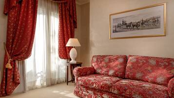 Una habitación con un sofá rojo estampado, un cuadro histórico enmarco y cortinas rojas con flecos.