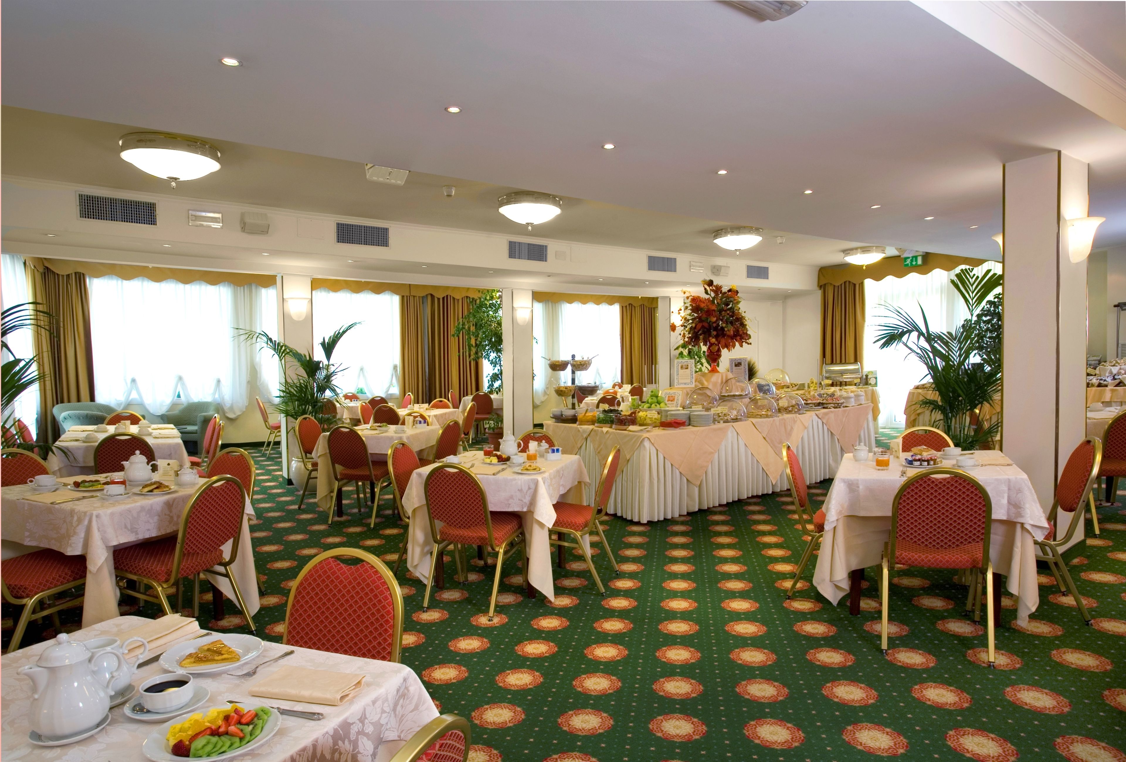 A hotel dining area with multiple tables and chairs, a buffet setup, and a green patterned carpet.