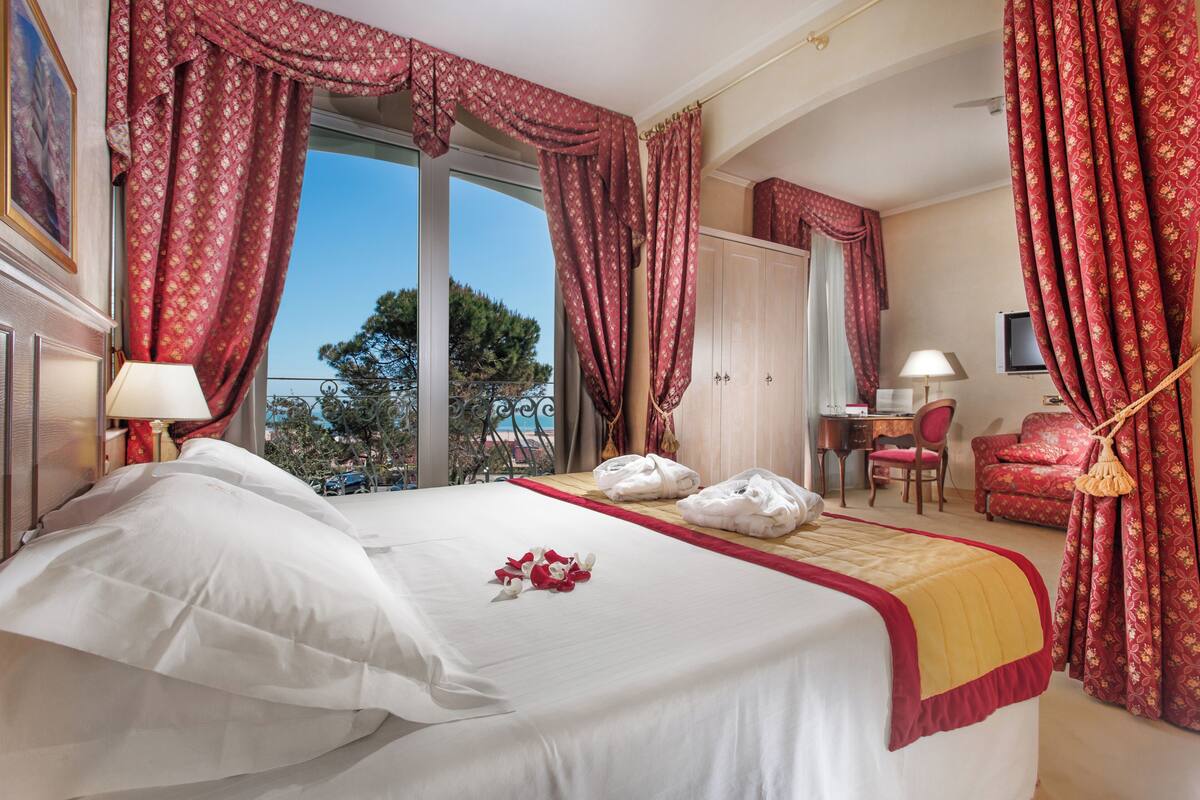 Ein Hotelzimmer mit einem großen Bett, einem Schreibtisch, einem Sessel und Blick ins Freie.