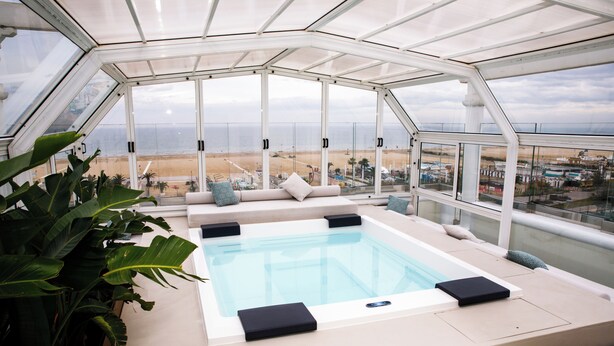 Piscina infinita en la azotea con barandilla de cristal, zona de estar con cojines y vistas a la playa y al paisaje urbano.