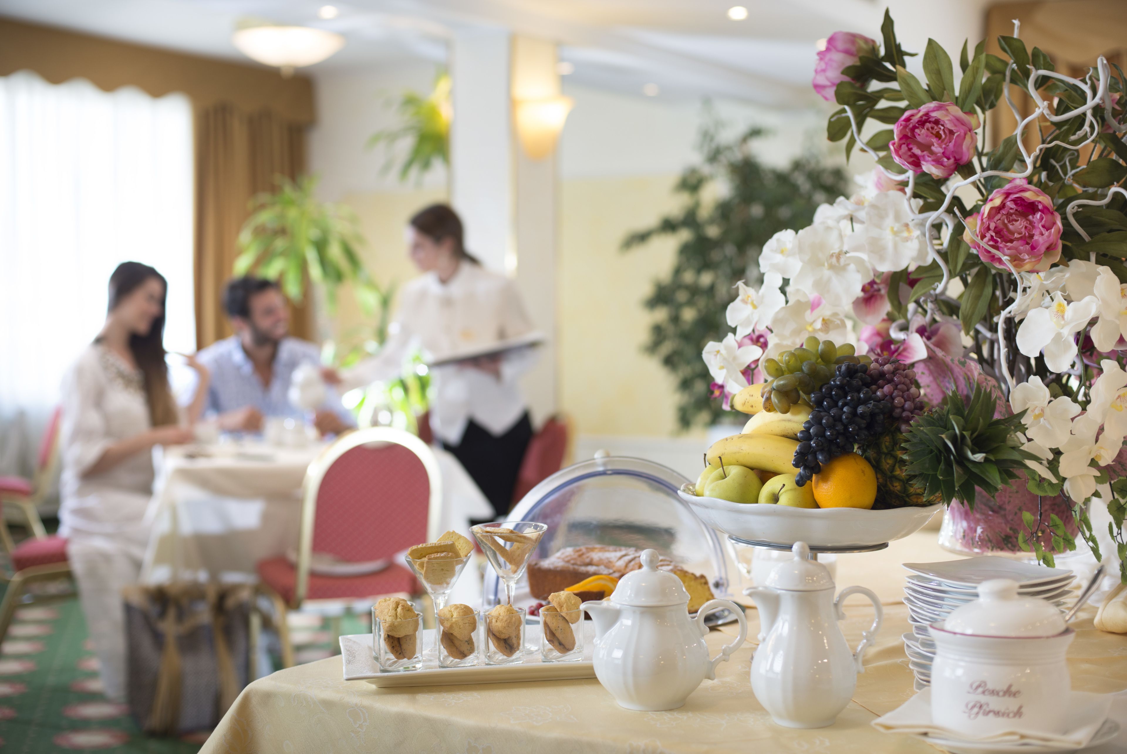 A dining table set with a fruit bowl, pastries, a pitcher, and a vase of flowers.