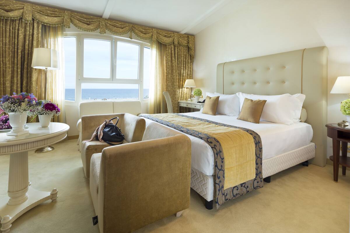 Ein Hotelzimmer mit einem großen Bett, einer Sitzecke mit Sessel und Tisch sowie Meerblick durch ein Fenster.