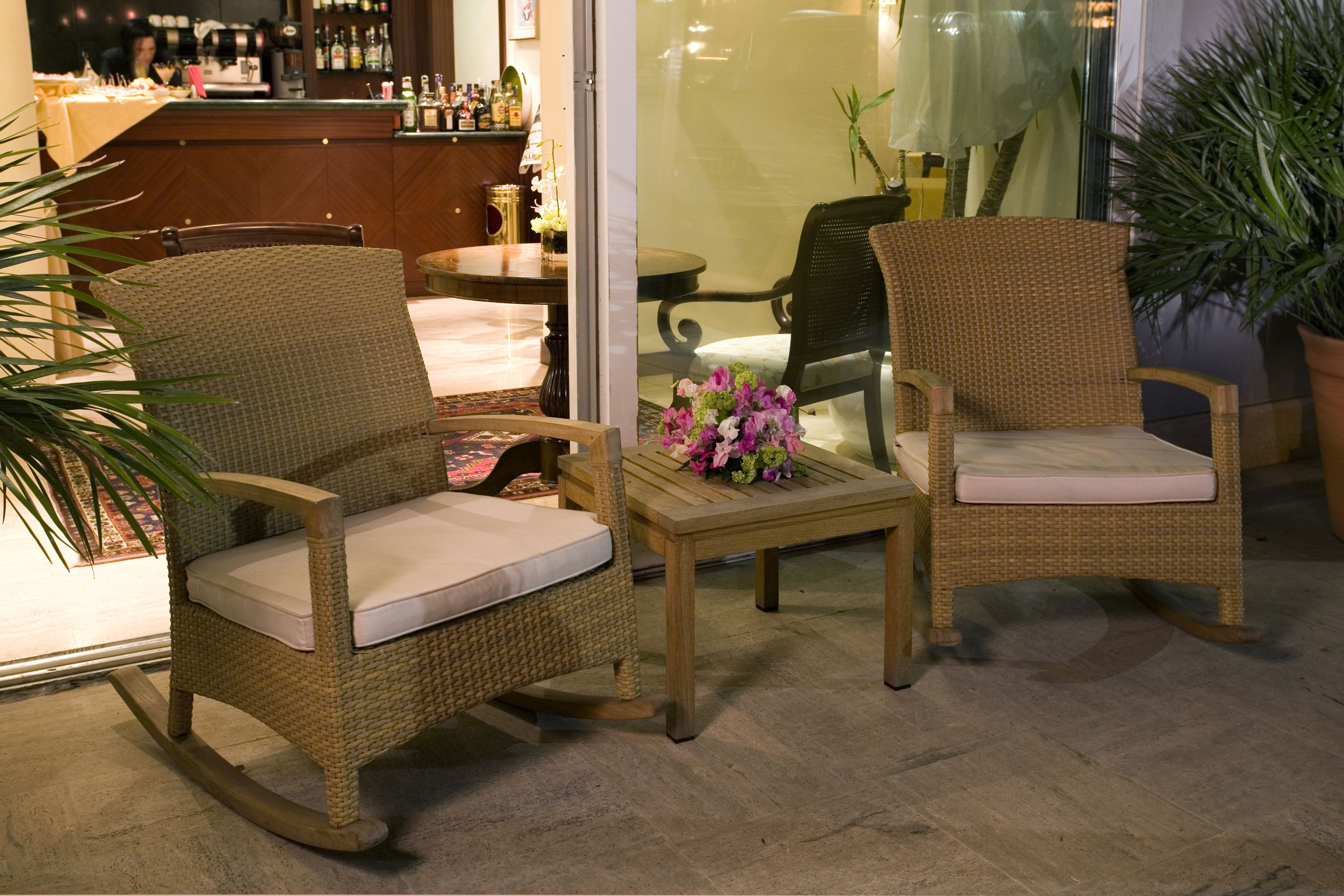 Wicker chairs with cushions, a small table with a vase of flowers, and a bar area in the background.