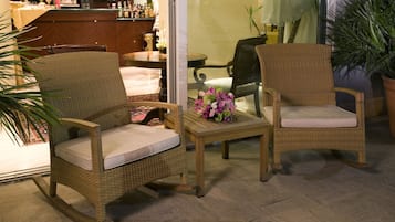 Wicker chairs with cushions, a small table with a vase of flowers, and a bar area in the background.