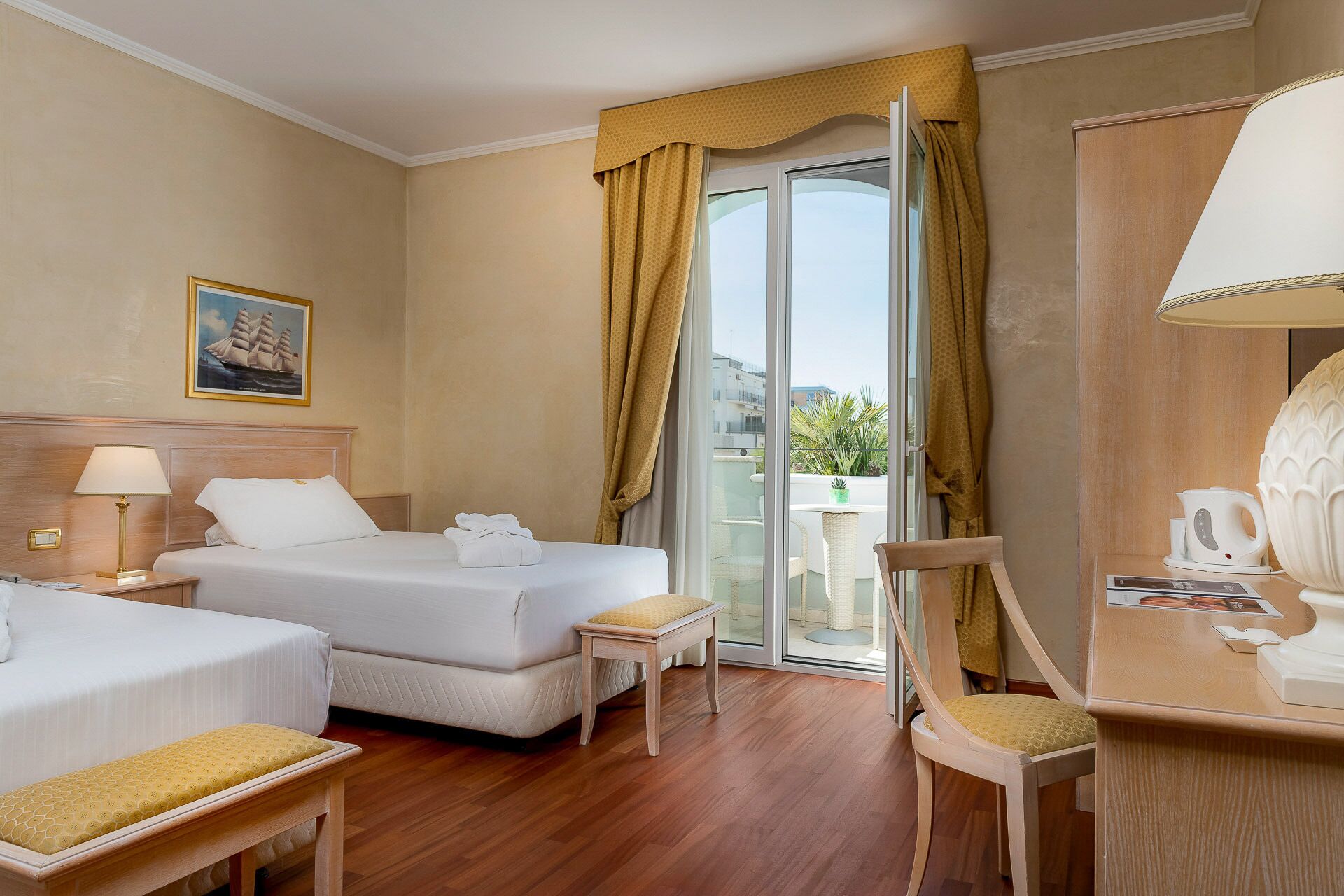 Habitación de hotel con dos camas, una zona de estar con una silla y una mesa, un balcón con cortinas amarillas y un cuadro en la pared.