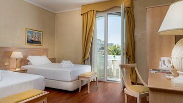 Habitación de hotel con dos camas, una zona de estar con una silla y una mesa, un balcón con cortinas amarillas y un cuadro en la pared.