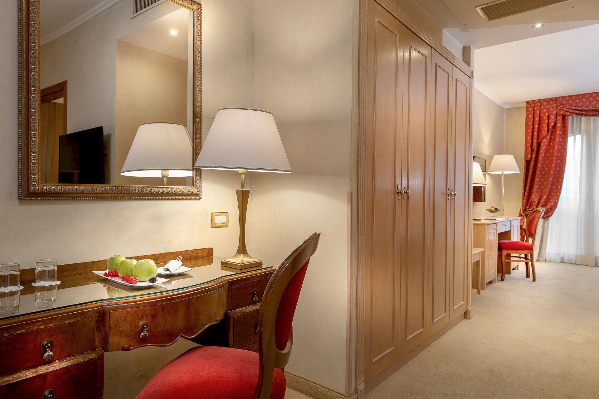 A hotel room with a wooden desk, two lamps, a mirror, a wardrobe, and a dining area with a chair and table.