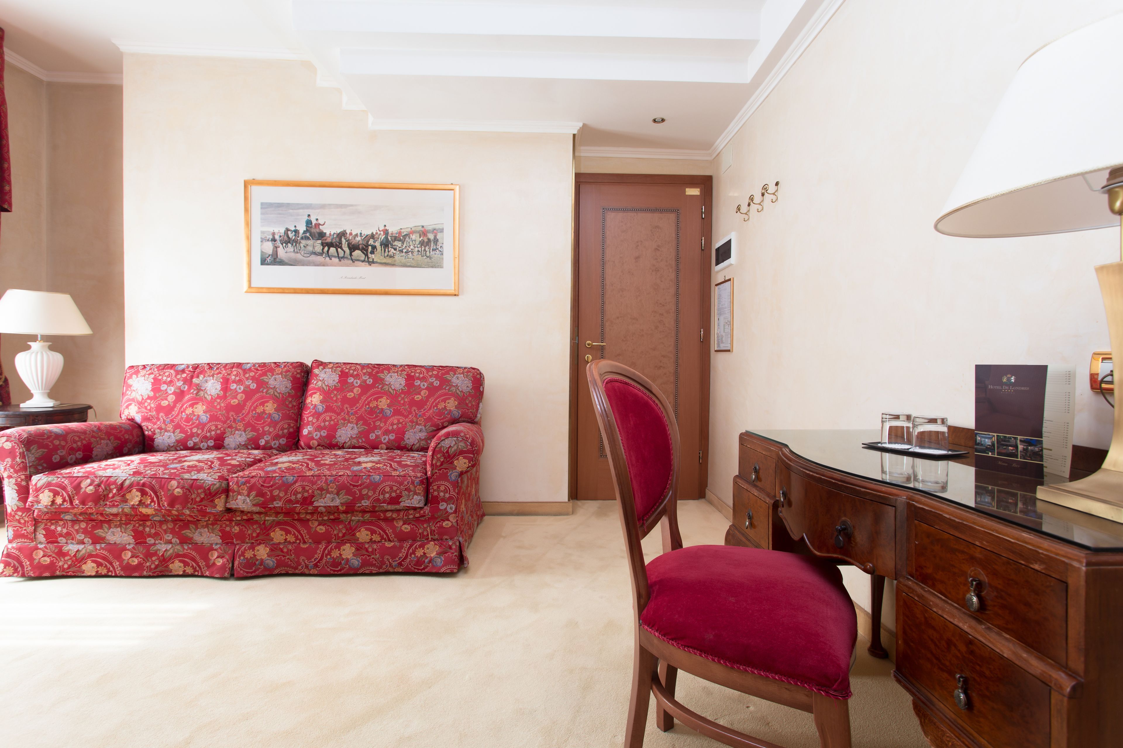 Una habitación con un sofá estampado con motivos florales, un escritorio de madera con cajones, una silla, un cuadro enmarcado en la pared y una puerta.