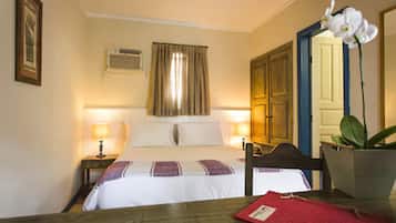 Een hotelkamer met een bed, nachtlampjes, een houten stoel, een tafel, een rode map, een plant in pot en een spiegel.